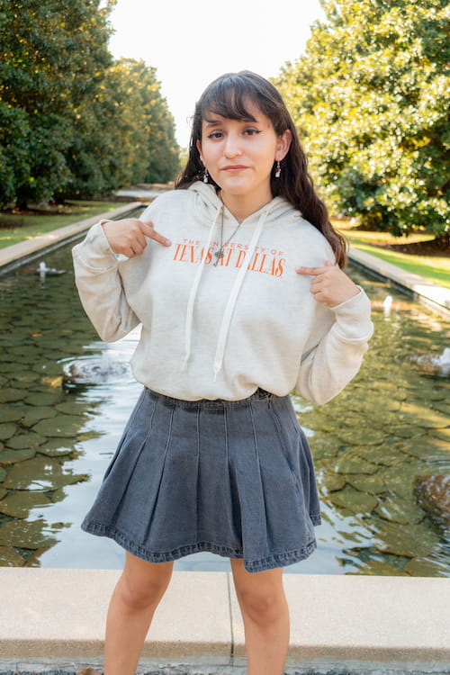 Female wearing hoodie, gesturing towards the University of Texas at Dallas lettering on the front.