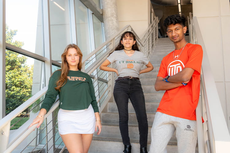 Three standing figures on a staircase, each modeling UT Dallas branded apparel.
